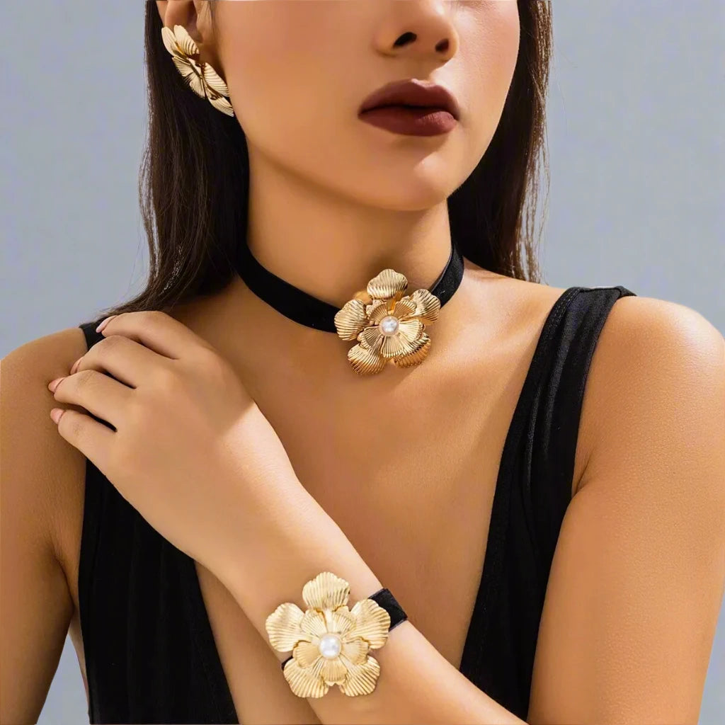 Striking jewelry set featuring bold gold-tone floral designs, including statement earrings, a choker, and a bracelet, each adorned with a detailed flower motif and a pearl center, creating a glamorous and cohesive look.