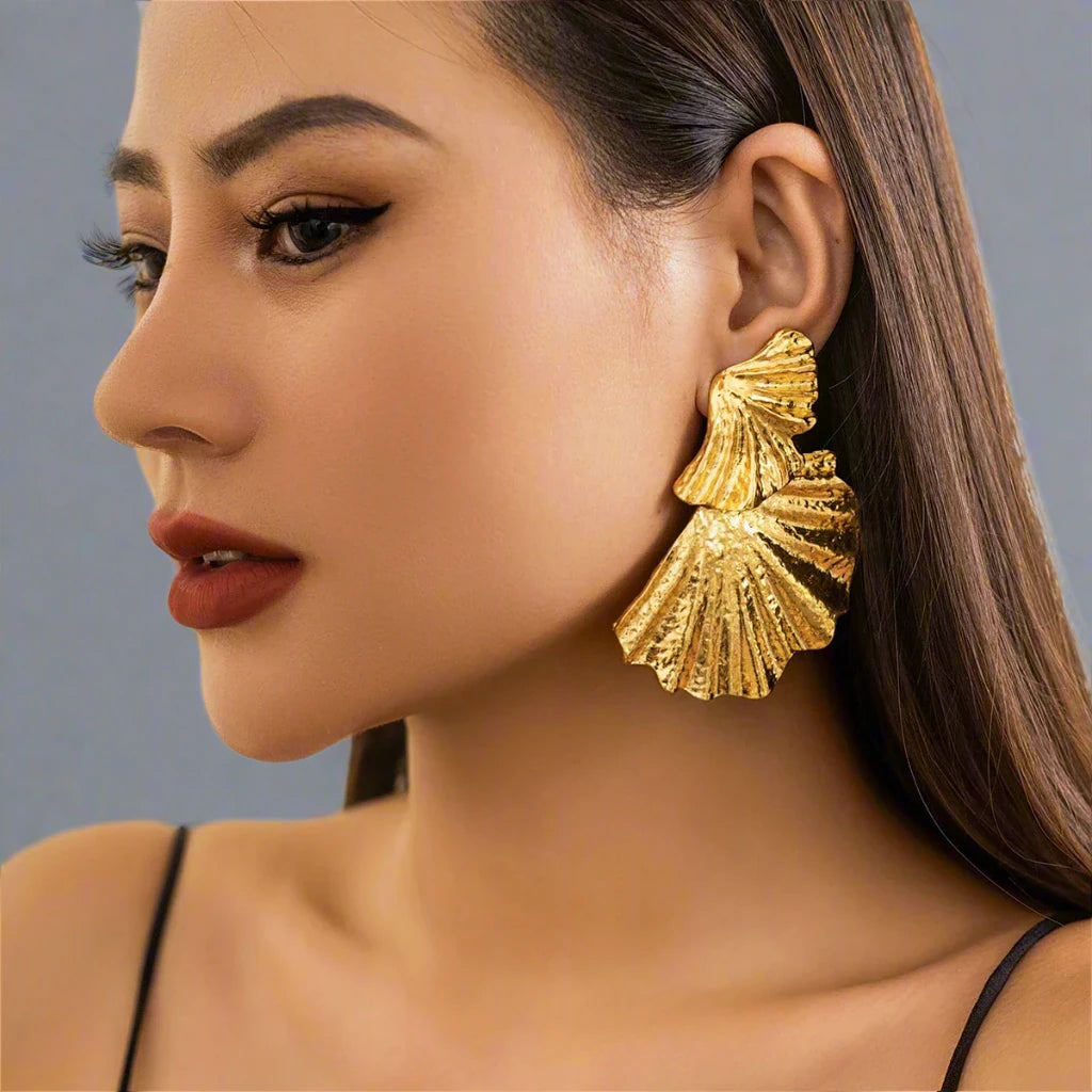 Gold-tone statement earring with a textured, fan-like design. The earring features multiple layered, shell-inspired shapes with intricate ridged details, adding an elegant yet bold look. The matte finish and unique structure give the earring a refined, sculptural aesthetic.