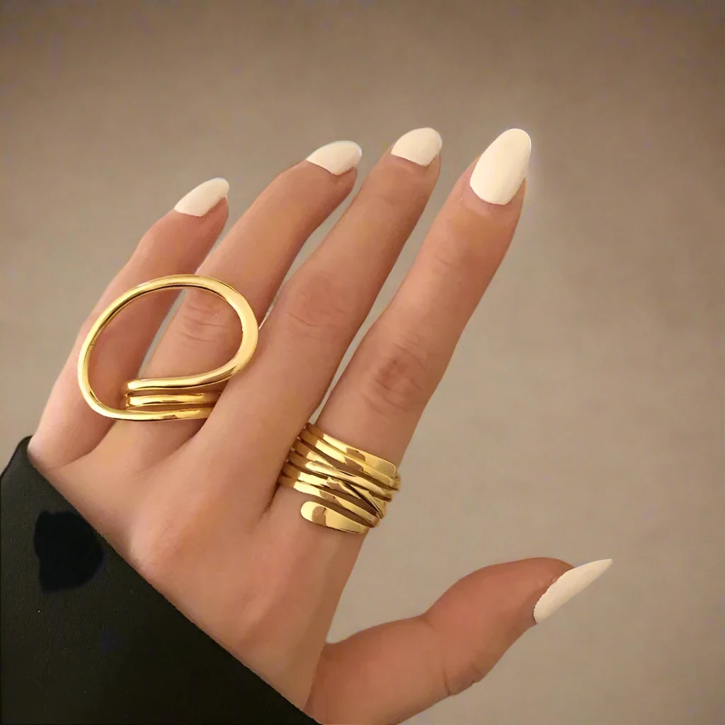 A set of two modern gold-tone rings featuring unique, abstract designs—one with a bold, open loop and the other with a layered, wrap-around style—creating a chic and artistic statement on the hand.