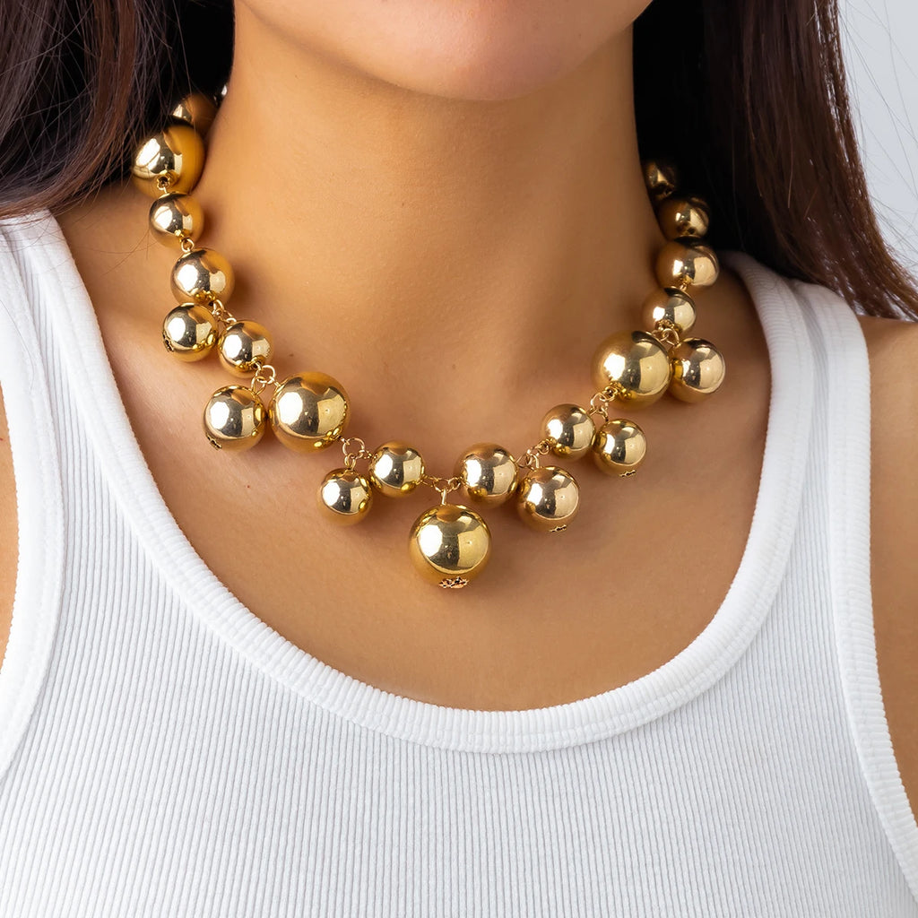 Bold gold-tone statement necklace with large, glossy metallic beads of varying sizes. The necklace adds a glamorous and chic touch to her simple white tank top.







