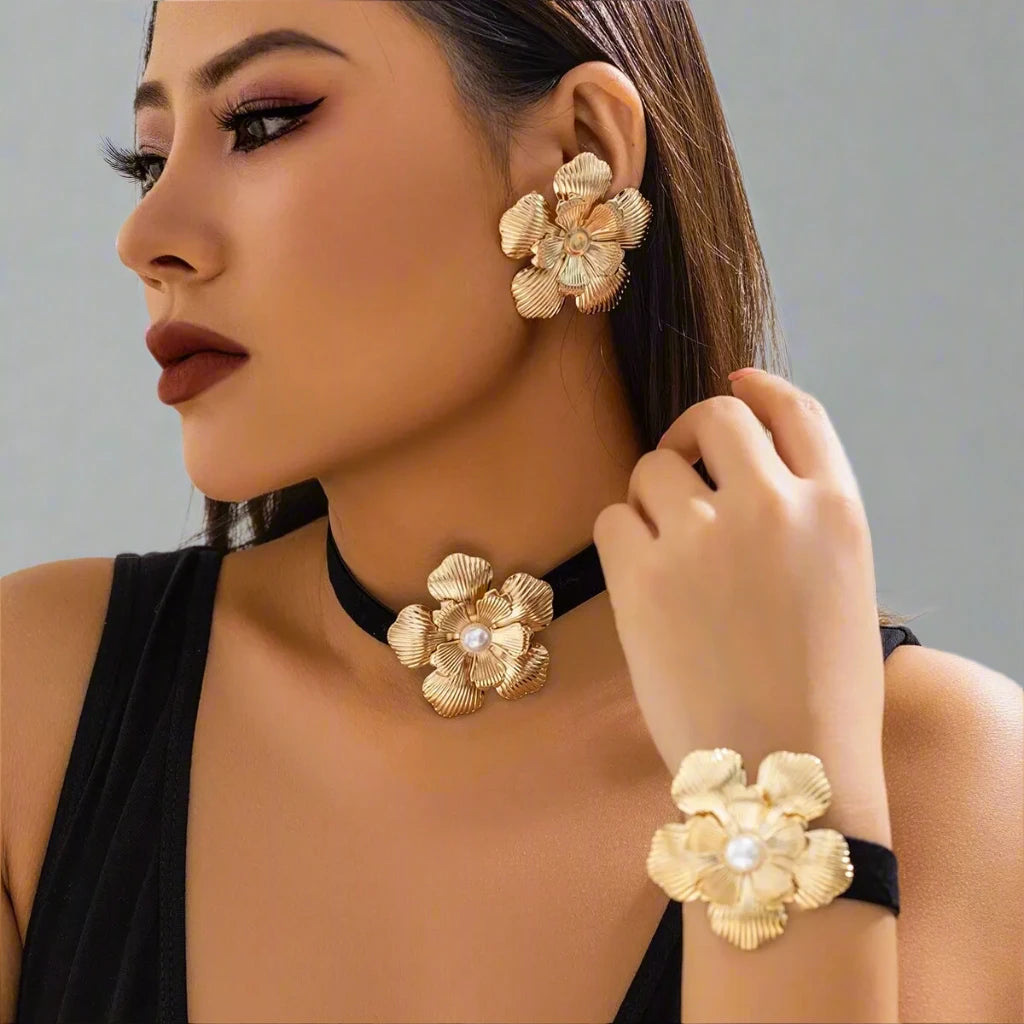 Striking jewelry set featuring bold gold-tone floral designs, including statement earrings, a choker, and a bracelet, each adorned with a detailed flower motif and a pearl center, creating a glamorous and cohesive look.