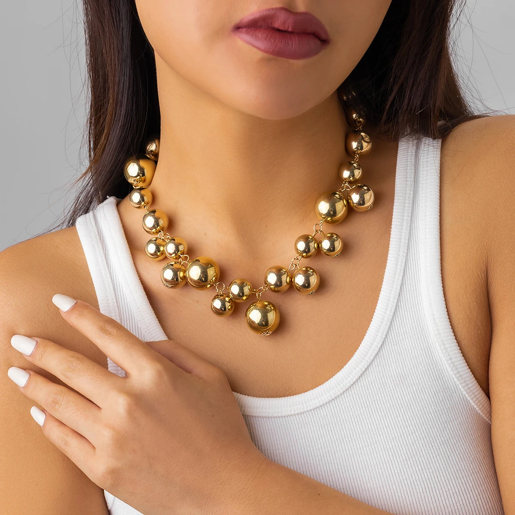 Bold gold-tone statement necklace with large, glossy metallic beads of varying sizes. The necklace adds a glamorous and chic touch to her simple white tank top.







