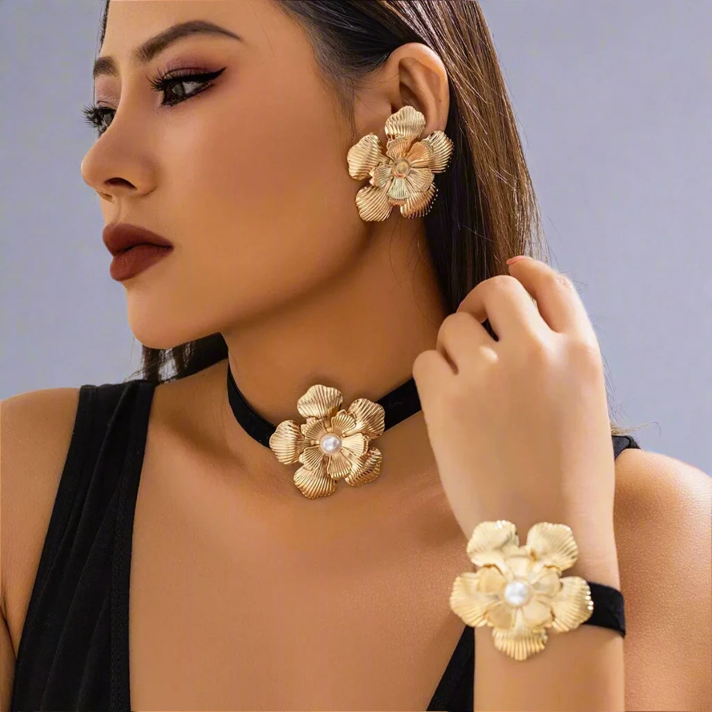 Striking jewelry set featuring bold gold-tone floral designs, including statement earrings, a choker, and a bracelet, each adorned with a detailed flower motif and a pearl center, creating a glamorous and cohesive look.