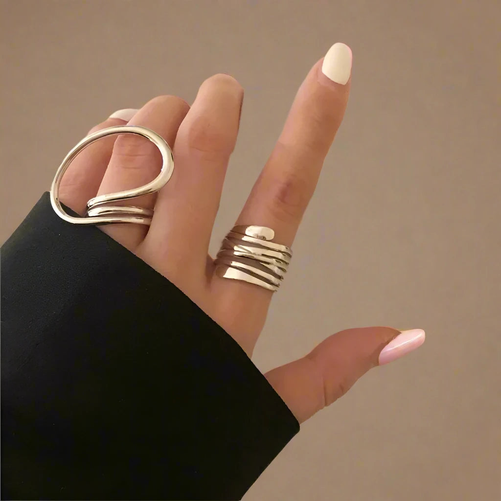 A set of two modern silver-tone rings featuring unique, abstract designs—one with a bold, open loop and the other with a layered, wrap-around style—creating a chic and artistic statement on the hand.