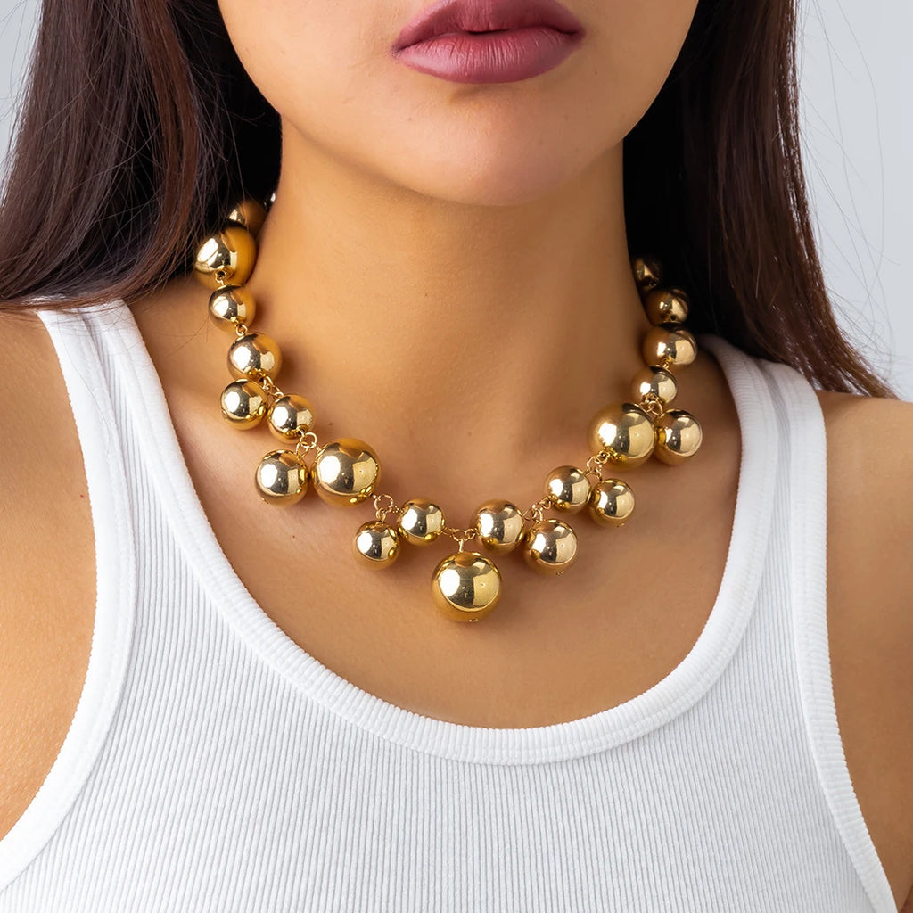 Bold gold-tone statement necklace with large, glossy metallic beads of varying sizes. The necklace adds a glamorous and chic touch to her simple white tank top.







