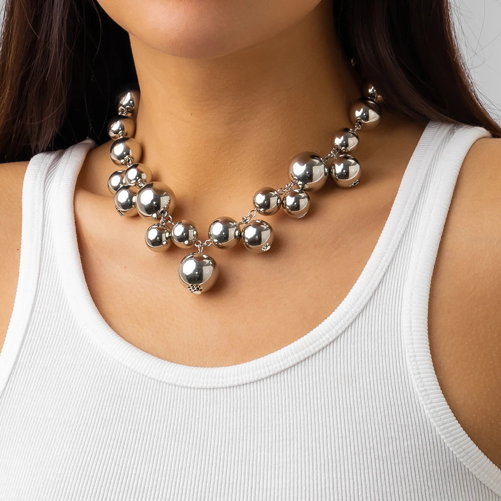 Bold Silver-tone statement necklace with large, glossy metallic beads of varying sizes. The necklace adds a glamorous and chic touch to her simple white tank top.







