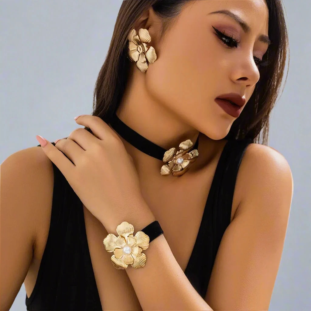 Striking jewelry set featuring bold gold-tone floral designs, including statement earrings, a choker, and a bracelet, each adorned with a detailed flower motif and a pearl center, creating a glamorous and cohesive look.