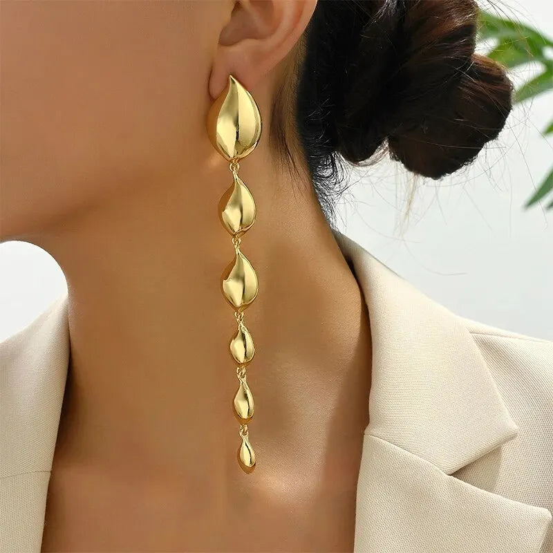 Gold-tone drop earrings with a sleek, elongated teardrop design. The earrings consist of multiple linked teardrop shapes, creating a cascading effect. The shiny metallic finish adds a modern and sophisticated touch to her neutral-colored outfit.







