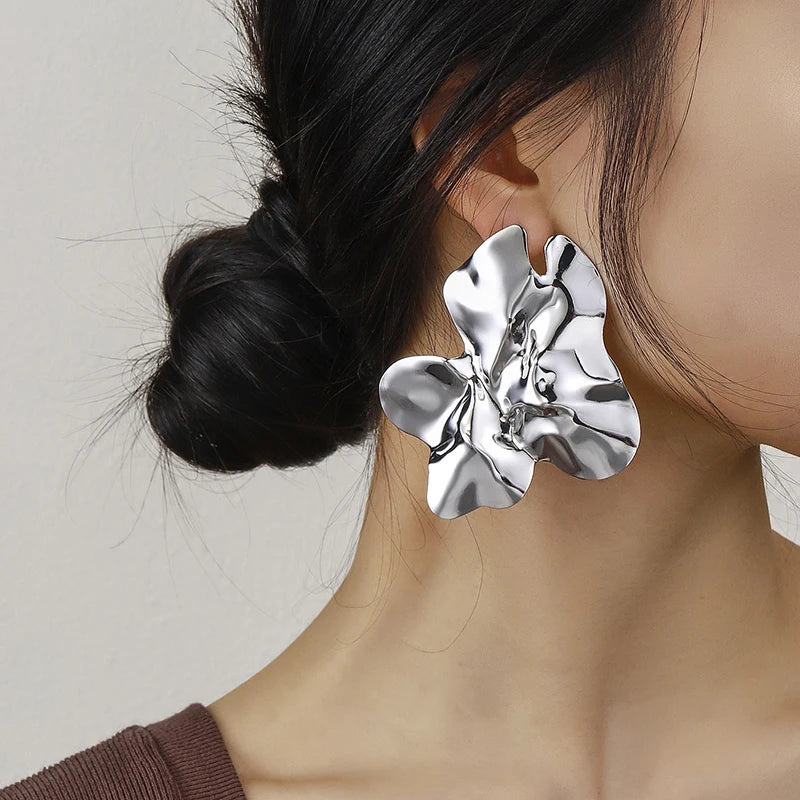 Large, silver-tone Floral sculptural earring. The earring has a fluid, abstract flower-like design with a shiny, reflective surface, adding a bold, artistic touch to her look.